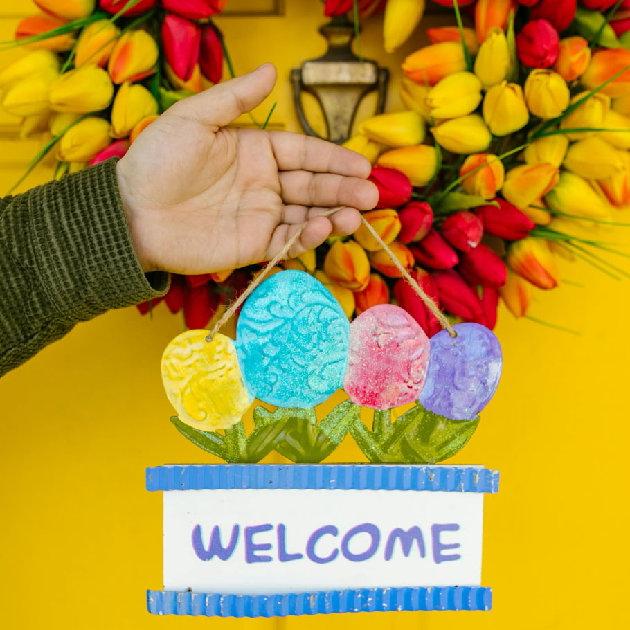A hand holding a vibrant Easter welcome sign against a tulip wreath on a yellow door.