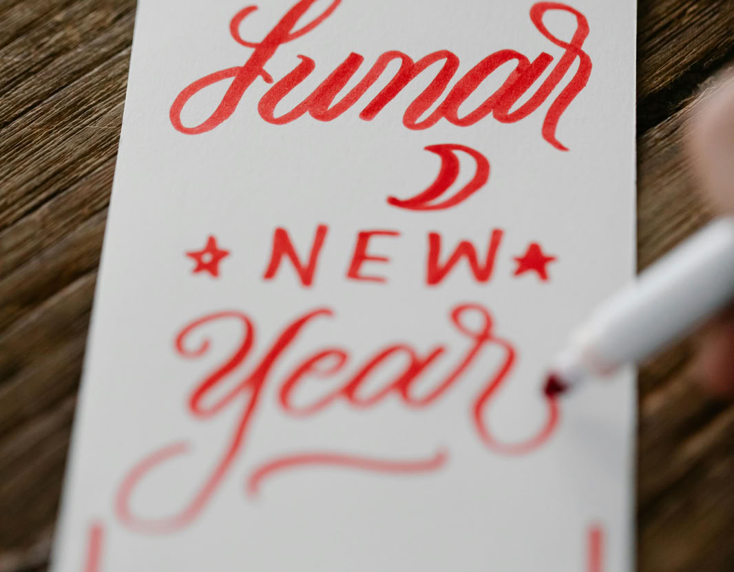 Close-up of a handwritten Lunar New Year card in red calligraphy on a wooden table.