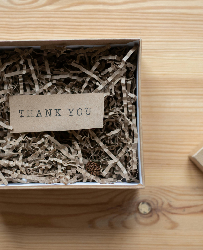 Top view of opened carton present box with small postcard with Thank You inscription and paper stuffing on wooden table